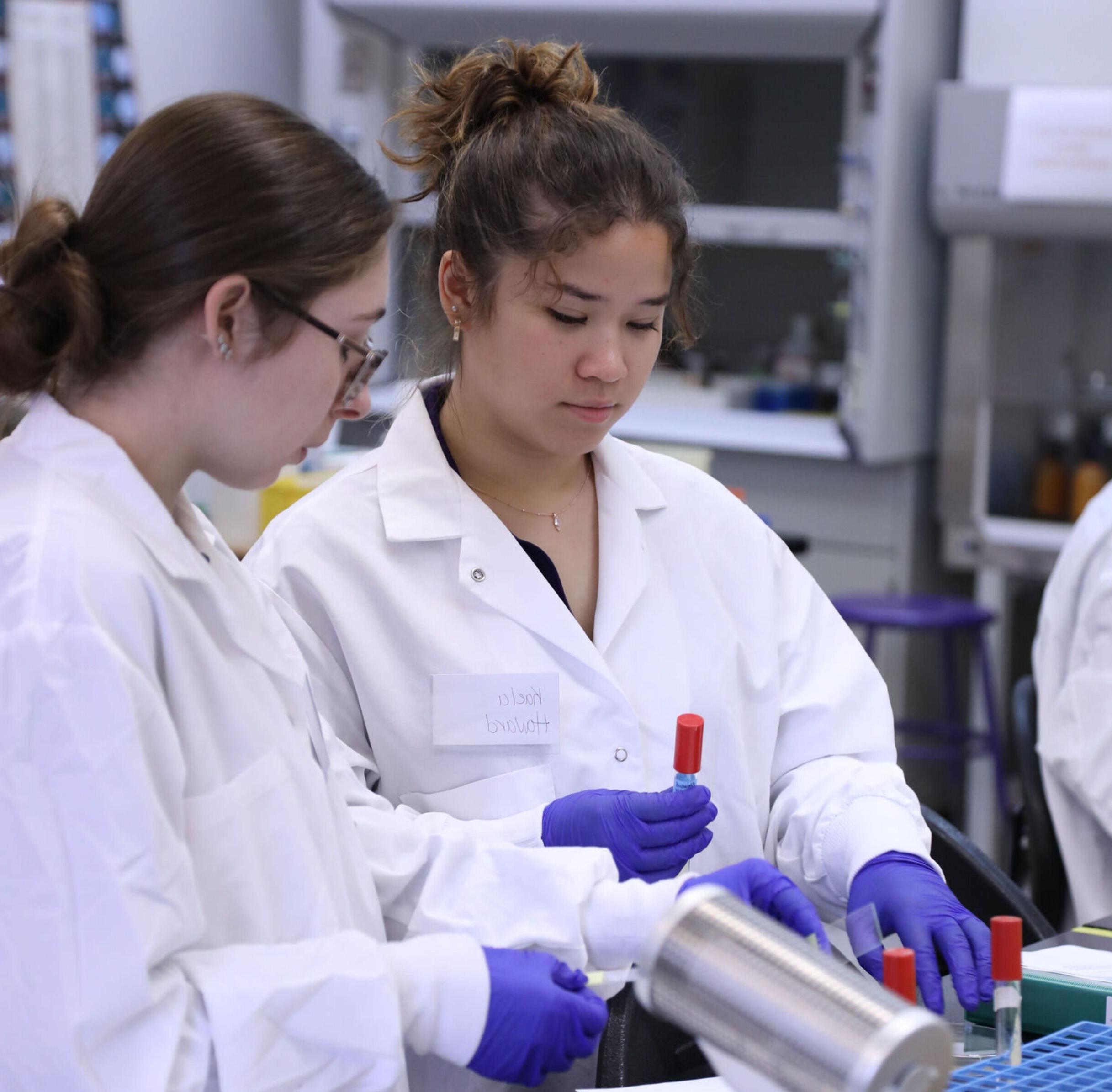 两个穿着实验室大褂的女学生在实验室里摆弄小瓶